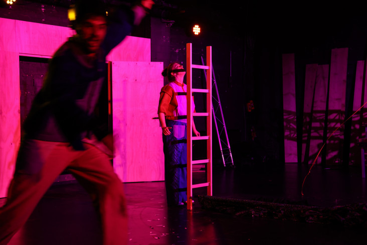 Die Bühne ist in pinkes Licht getaucht. Vorn läuft ein Schauspieler. Hinten steht eine Schauspielerin mit einer Leiter. Die Bühne ist in pinkes Licht getaucht. Vorn läuft ein Schauspieler. Hinten steht eine Schauspielerin mit einer Leiter.