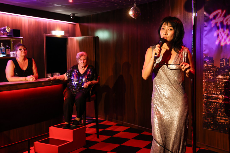 An einer Bar sitzt eine Frau auf einem Stuhl. Eine andere steht hinter der Bar. Beide blicken auf eine Performerin. Sie hält ein Mikro in der Hand und singt. An einer Bar sitzt eine Frau auf einem Stuhl. Eine andere steht hinter der Bar. Beide blicken auf eine Performerin. Sie hält ein Mikro in der Hand und singt.