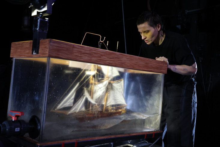 Eine Schauspielerin schaut in einen Glaskasten, in dem ein großes Segelschiff schwimmt. Eine Schauspielerin schaut in einen Glaskasten, in dem ein großes Segelschiff schwimmt.
