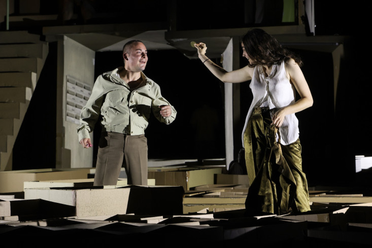 Die Bühne ist mit Umzugskartons bedeckt. In einem Karton steht eine Schauspielerin. Sie hält eine große Münze in der Hand. Ihr gegenüber steht ein Schauspieler. Beide lachen. Die Bühne ist mit Umzugskartons bedeckt. In einem Karton steht eine Schauspielerin. Sie hält eine große Münze in der Hand. Ihr gegenüber steht ein Schauspieler. Beide lachen.