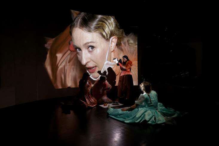 Vier Schauspielerinnen sind auf der Bühne, eine filmt das Gesicht einer anderen. Deren Gesicht ist groß auf eine Wand projeziert. Vier Schauspielerinnen sind auf der Bühne, eine filmt das Gesicht einer anderen. Deren Gesicht ist groß auf eine Wand projeziert.
