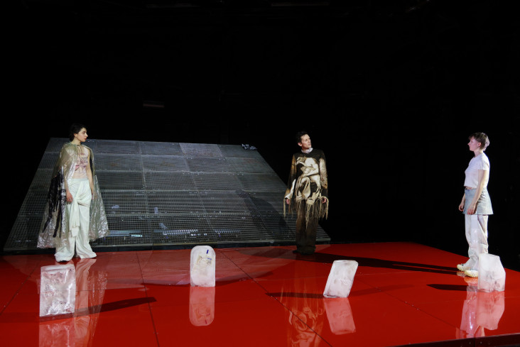 Der Bühnenboden ist leuchtend rot. Drei Personen stehen mit Abstand zueinander auf der Bühne. Vor ihnen stehen Eisblöcke.