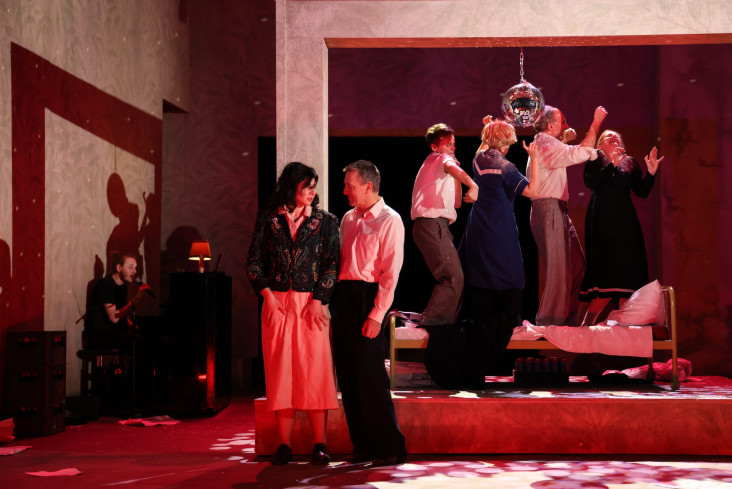 Ein Raum ist in rotes Licht getaucht. Im Hintergrund tanzen mehrere Menschen unter einer Diskokugel. Vorn blicken sich eine Schauspielerin und ein Schauspieler an. Ein Raum ist in rotes Licht getaucht. Im Hintergrund tanzen mehrere Menschen unter einer Diskokugel. Vorn blicken sich eine Schauspielerin und ein Schauspieler an.