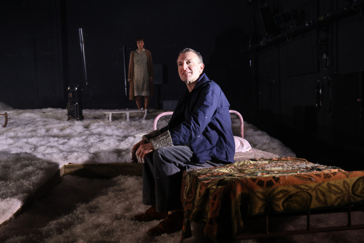 Ein Schauspieler sitzt allein auf einem Bett. Weit hinter ihm auf der Bühne steht eine Schauspielerin. Ein Schauspieler sitzt allein auf einem Bett. Weit hinter ihm auf der Bühne steht eine Schauspielerin.