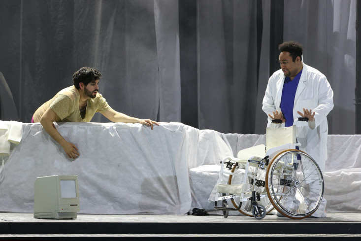 Ein Sänger stützt sich auf einem weißen Möbelstück auf. Er schaut zu einem anderen Sänger, der einen Krankenhauskittel trägt. Er schiebt einen Rollstuhl.