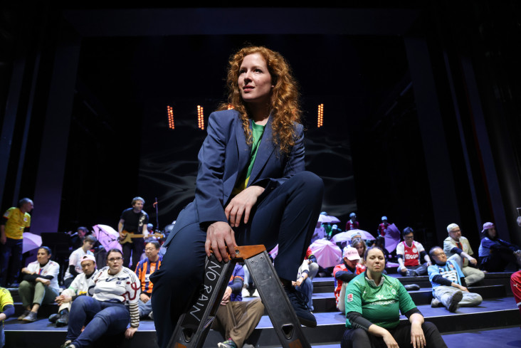 Eine Schauspielerin sitzt auf einer Leiter. Hinter ihr sitzen viele Personen verteilt auf der Bühne. Eine Schauspielerin sitzt auf einer Leiter. Hinter ihr sitzen viele Personen verteilt auf der Bühne.