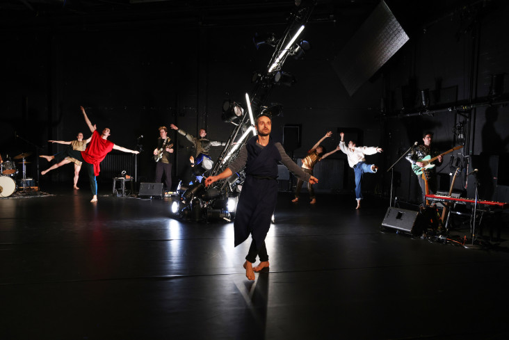 Über die Bühne verteilt stehen einzelne Musiker mit ihren Instrumenten. In der Mitte der Bühne gibt es eine Installation aus Scheinwerfern. Um die herum tanzen sechs Personen.  Über die Bühne verteilt stehen einzelne Musiker mit ihren Instrumenten. In der Mitte der Bühne gibt es eine Installation aus Scheinwerfern. Um die herum tanzen sechs Personen.