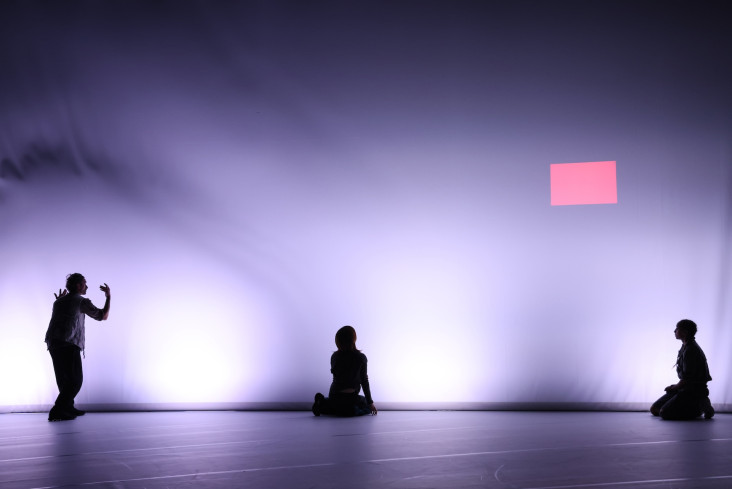 Im Hintergrund der Bühne ist eine riesige Leinwand gespannt. Sie wird von hinten beleuchtet. Vor ihr sind drei Personen. Sie haben einen großen Abstand zueinander. Im Hintergrund der Bühne ist eine riesige Leinwand gespannt. Sie wird von hinten beleuchtet. Vor ihr sind drei Personen. Sie haben einen großen Abstand zueinander.