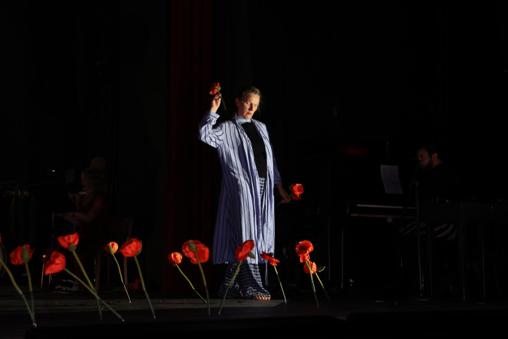 Eine Schauspielerin steht auf einer dunklen Bühne. In der Hand hält sie eine Mohnblume. Vor ihr stehen viele Mohnenblumen. 