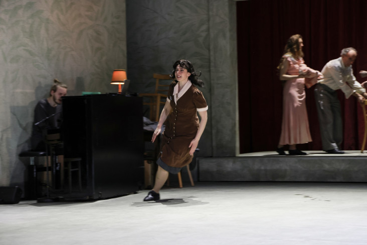 Eine Schauspielerin rennt über die Bühne. Sie trägt ein braunes Kleid. Hinter ihr sitzt ein Mann an einem Klavier. Eine Schauspielerin rennt über die Bühne. Sie trägt ein braunes Kleid. Hinter ihr sitzt ein Mann an einem Klavier.