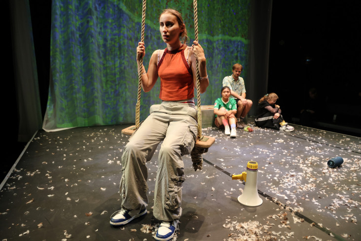 Auf der Schaukel vorn sitzt eine Schauspielerin. Hinten auf der Bühne sitzen drei Personen eng beieinander. Sie sehen alle zu der Person auf der Schaukel hin. Auf der Schaukel vorn sitzt eine Schauspielerin. Hinten auf der Bühne sitzen drei Personen eng beieinander. Sie sehen alle zu der Person auf der Schaukel hin.