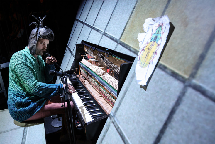 Ein Schauspieler sitzt an einem Klavier. Er trägt eine Ohrenmütze auf dem Kopf. An der Mütze sind zwei Fühler. Ein Schauspieler sitzt an einem Klavier. Er trägt eine Ohrenmütze auf dem Kopf. An der Mütze sind zwei Fühler.