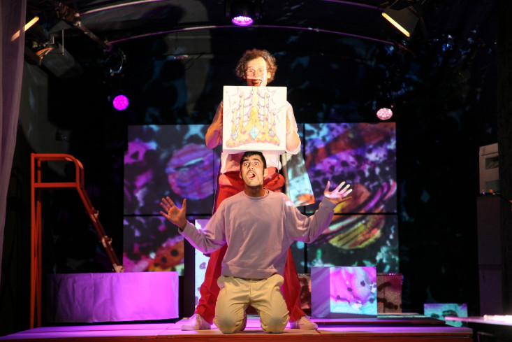 Ein Schauspieler kniet auf dem Boden. Hinter ihm steht ein Sänger. Der hält eine Zeichnung über den Kopf des Knienden. Hinter ihnen ist alles bunt erleuchtet. Ein Schauspieler kniet auf dem Boden. Hinter ihm steht ein Sänger. Der hält eine Zeichnung über den Kopf des Knienden. Hinter ihnen ist alles bunt erleuchtet.