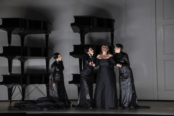 Vier Sängerinnen in schwarzen Kleidern stehen auf der Bühne. Zwei von ihnen stützen eine. Hinter ihnen sind jeweils drei Flügel übereinandergebaut. 
