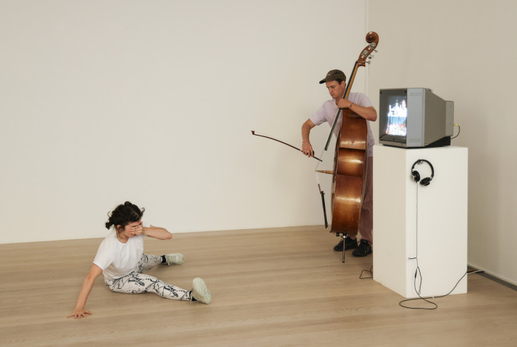 Neben einem Bildschirm steht ein Musiker. Er spielt Bass. Auf dem Boden vor ihm sitzt eine Tänzerin. Sie ist mitten in einer Drehung. Neben einem Bildschirm steht ein Musiker. Er spielt Bass. Auf dem Boden vor ihm sitzt eine Tänzerin. Sie ist mitten in einer Drehung.