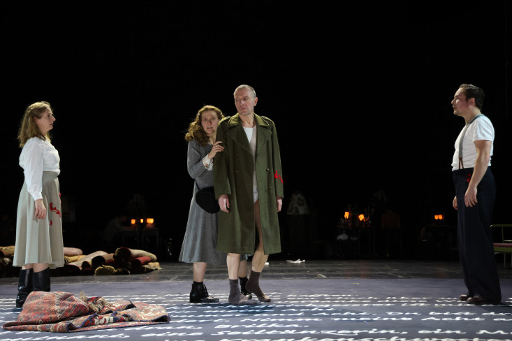 Eine Schauspielerin hält einen Schauspieler im Militärmantel an den Schultern. Er trägt keine Hose. Links und rechts von ihnen stehen zwei Personen und schauen sie an. Eine Schauspielerin hält einen Schauspieler im Militärmantel an den Schultern. Er trägt keine Hose. Links und rechts von ihnen stehen zwei Personen und schauen sie an.