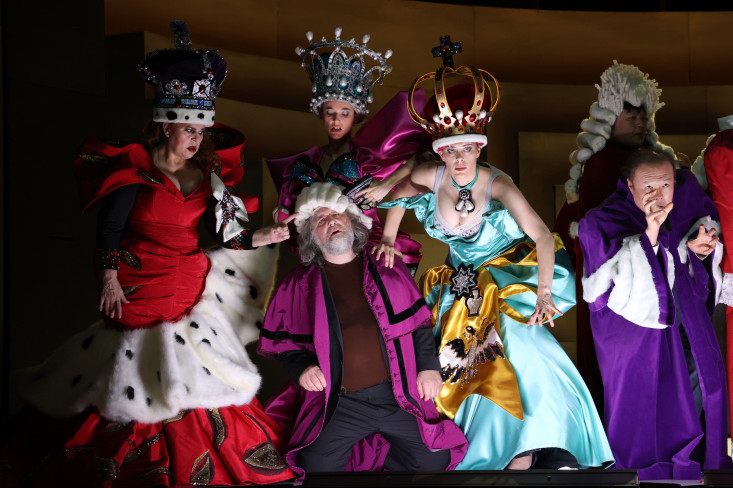 Ein Mann kniet auf der Bühne. Er ist umgeben von drei Frauen in opulenten Königinnenkostümen.