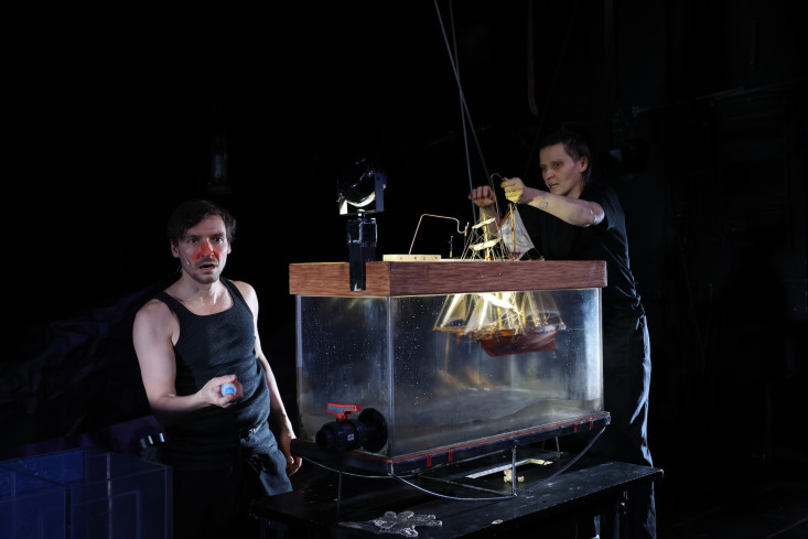 Ein Schauspieler spricht. Eine Schauspielerin senkt ein großes Segelschiff in einen Glaskasten. Ein Schauspieler spricht. Eine Schauspielerin senkt ein großes Segelschiff in einen Glaskasten.