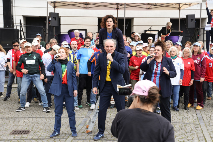 Vier Personen stehen im Vordergrund. Alle halten Mikrofone in den Händen. Hinter ihnen stehen sehr viele Menschen. Alle tragen Fußballtrikots. Vier Personen stehen im Vordergrund. Alle halten Mikrofone in den Händen. Hinter ihnen stehen sehr viele Menschen. Alle tragen Fußballtrikots.