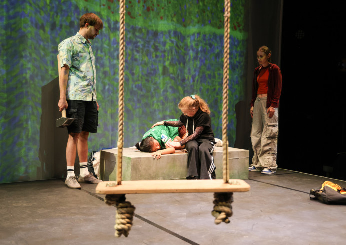 Vorn im Bild hängt eine Schaukel. Hinten liegt eine Person auf einem Kasten. Eine andere sitzt neben ihr und legt ihr den Arm auf den Rücken. Zwei andere schauen zu. Vorn im Bild hängt eine Schaukel. Hinten liegt eine Person auf einem Kasten. Eine andere sitzt neben ihr und legt ihr den Arm auf den Rücken. Zwei andere schauen zu.