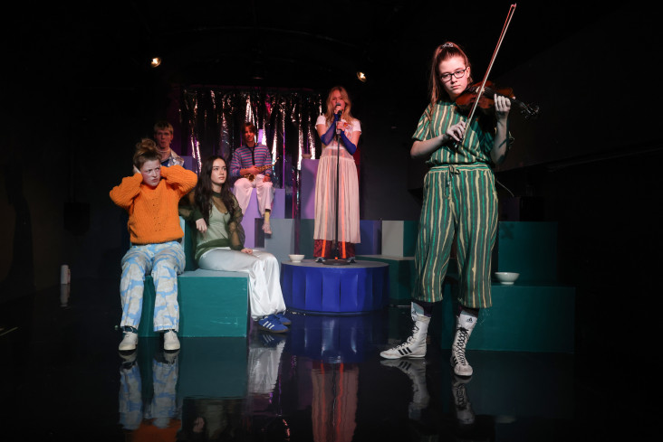 Sechs junge Akteure sind auf der Bühne. Eine singt, eine spielt Geige. Eine hält sich die Ohren zu. Sechs junge Akteure sind auf der Bühne. Eine singt, eine spielt Geige. Eine hält sich die Ohren zu.