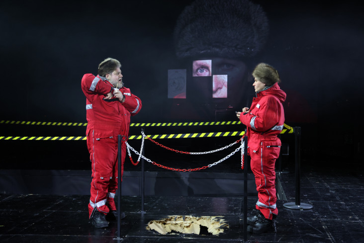 Zwei Personen in roten Anzügen stehen sich gegenüber. In der Mitte zwischen ihnen ist ein Loch im Boden. Um das Loch herum ist eine Absperrung.