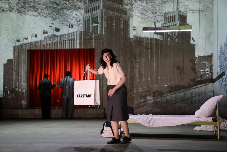 Eine Schauspielerin steht vorn auf der Bühne. Sie hält eine Tüte hoch. Auf der steht Karstadt. Im Hintergrund ist ein Bild der Bremer Innenstadt projiziert. Eine Schauspielerin steht vorn auf der Bühne. Sie hält eine Tüte hoch. Auf der steht Karstadt. Im Hintergrund ist ein Bild der Bremer Innenstadt projiziert.