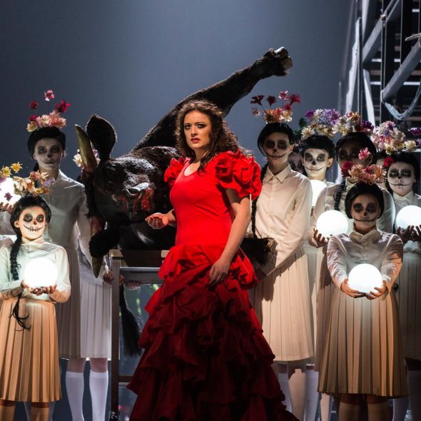 Carmen  | Zu den  Vorstellungen Auf dem Foto einen Szenenimpression aus CARMEN: Carmen in einem auffälligen roten Volantkleid, hinter und neben ihr Kinder in Kleidern, die den mexikanischen Día de los Muertos zitieren. Ein toter Stier liegt hinter Carmen auf der Seite.