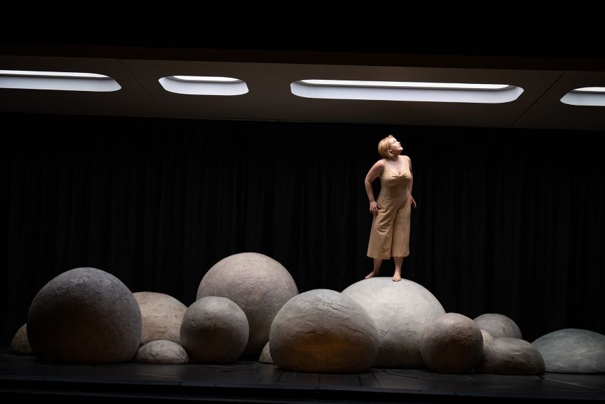 Ein dunkler Raum mit Leuchtfeldern an der Decke. Runde Felsen auf dem Boden. Eine Frau steht auf einem Felsen und blickt nach oben. © Sandra Then