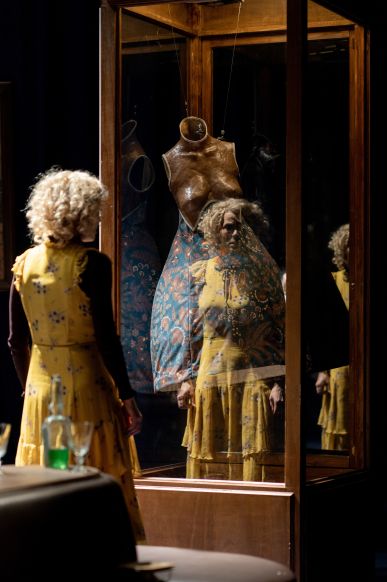 Das Bild zeigt eine blondgelockte mittelalte Frau in einem gelben Kleid mit braunem Unterziehpulli, die vor einer Holzvitrine steht. Sie betrachtet die darin stehende Büste mit langem blauen Kleid mit orangefarbenen Kreisen.  © Britt Schilling