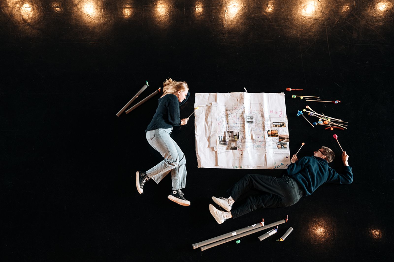 Die beiden Theaterpädagog*innen Charlie Rackwitz und Madeleine Kandschur liegen am Boden des Foyers zwischen einem Lageplan und kleinen Musikinstrumenten. Unter ihrer Leitung entsteht ein Opern-Parcours, den die Kinder des Kinderclubs inszenieren.